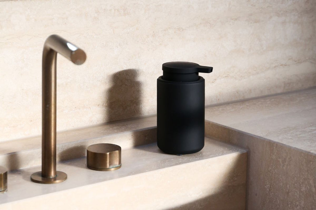 A matte black ARC soap dispenser sits on a beige travertine countertop beside a modern, curved brass faucet.
