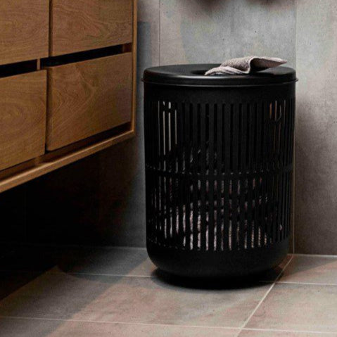 Natural light, perspective view of a black, cylindrical laundry basket on the floor in a concrete-walled bathroom with two towels hung above it.