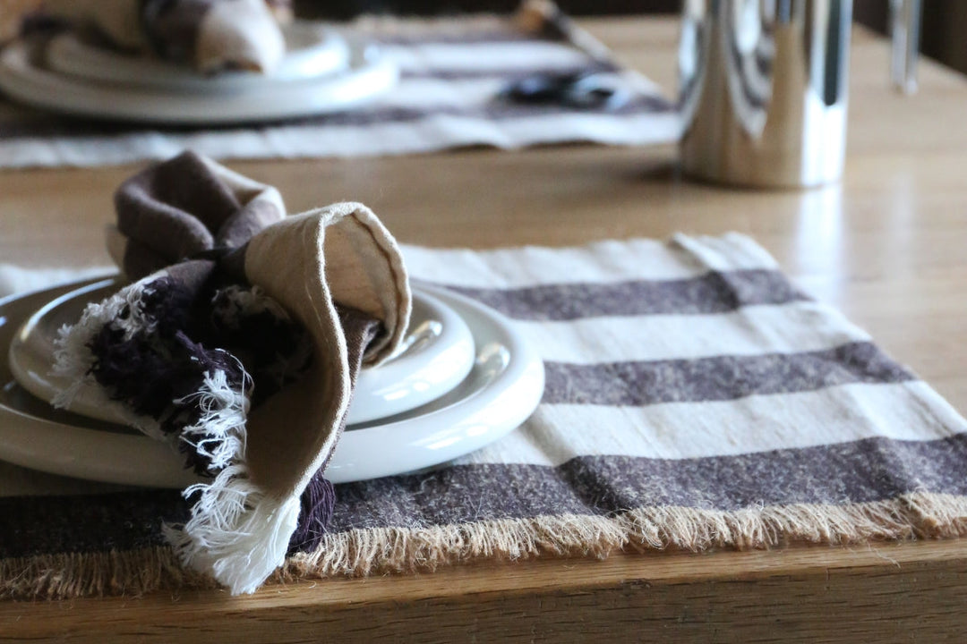 A close-up, angled shot of a set dinner table. In the foreground, a white ceramic plate sits on a woven, striped placemat with a fringed border. A brown and cream cloth napkin is artfully arranged on the plate. In the blurred background, another place set