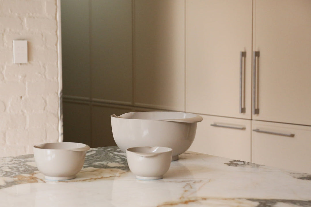 Three neutral-toned mixing bowls on a marble benchtop in a soft beige kitchen, styled with natural light and curved forms.