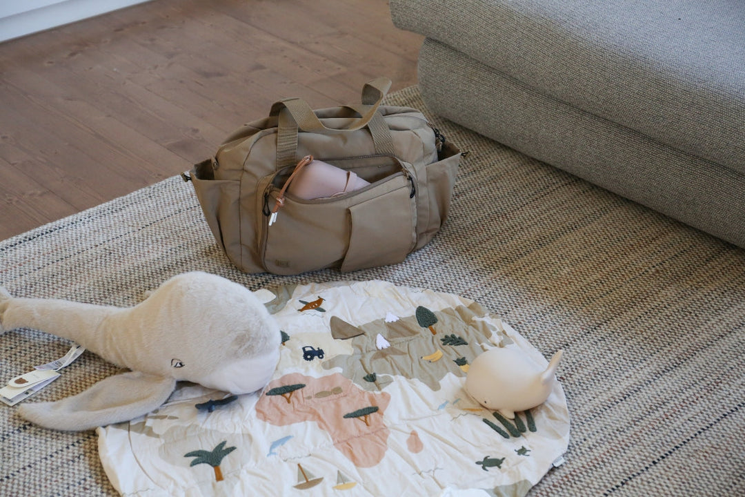Calming nursery setup featuring a world map play mat, plush whale toy, silicone night light, and structured nappy bag—all in soft neutral tones and styled on a woven rug. All products from LIEWOOD.