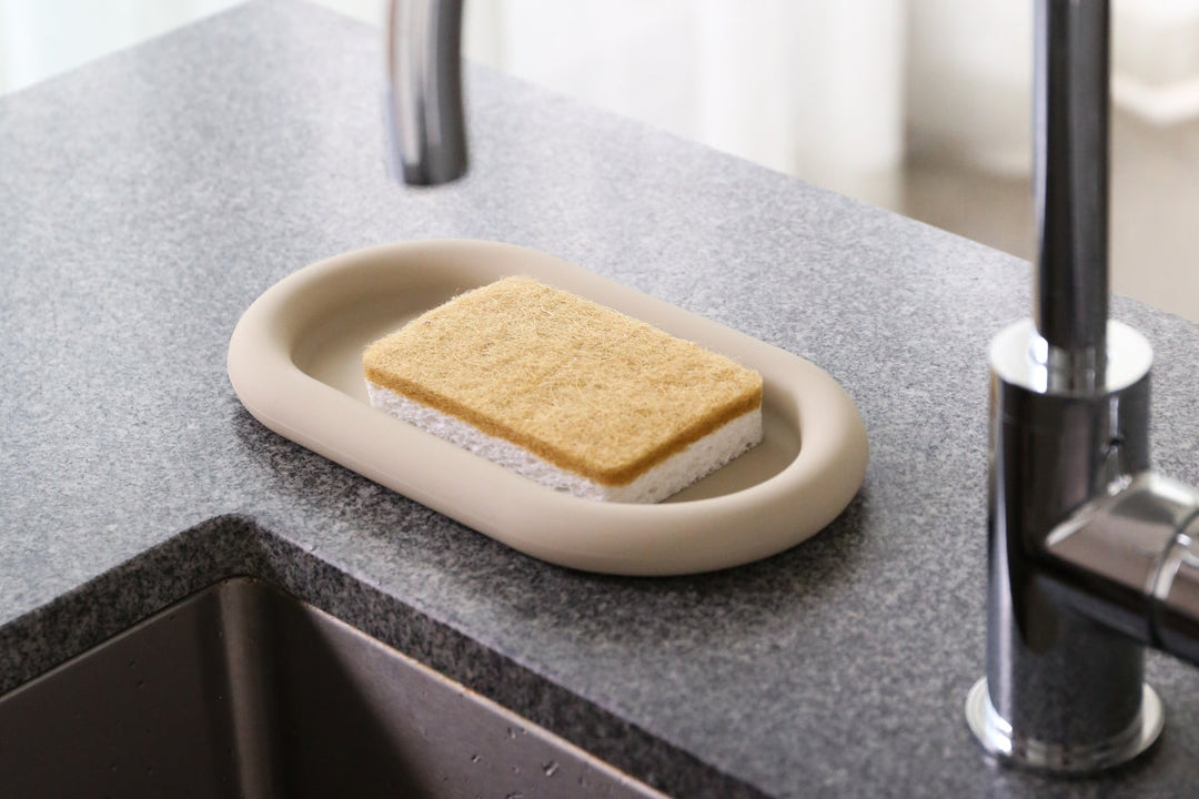 A natural cellulose and sisal scourer sponge resting on a neutral-toned sponge holder beside a kitchen sink. Discover the best eco-friendly sponges for cleaning in our latest guide.