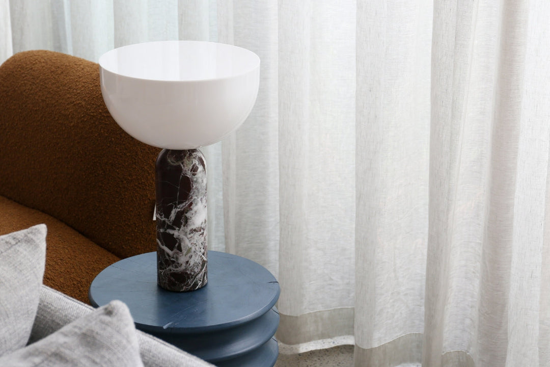 The New Works Kizu Lamp, featuring a striking Rosso Levanto marble base and soft, curved white shade, styled on a deep blue side table beside a textured brown sofa and sheer curtains.