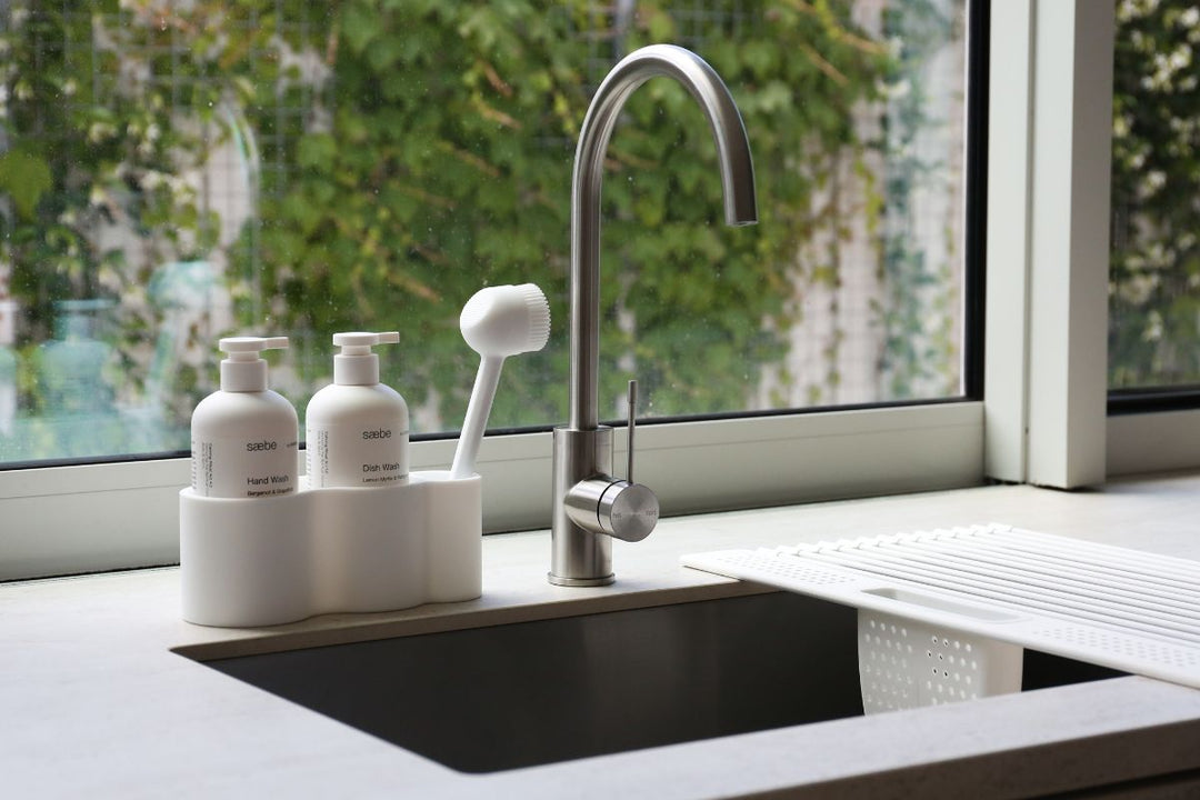 A calm kitchen sink area with a white sink-side organiser holding hand wash, dish wash and a dish brush beside a stainless steel tap and white drying rack, with greenery visible through the window.