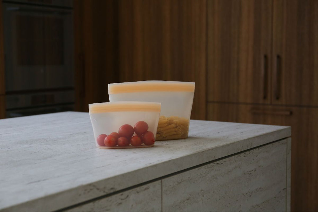 Two reusable silicone food pouches filled with cherry tomatoes and pasta on a stone kitchen bench, with timber cabinetry in the background.