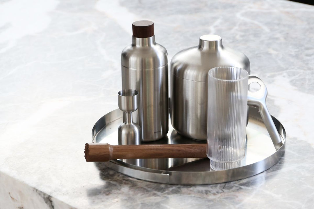 A stainless steel drinks tray on a marble bench holding a cocktail shaker, ice bucket, jigger, muddler, bar spoon and ribbed glass for a home drinks setup.