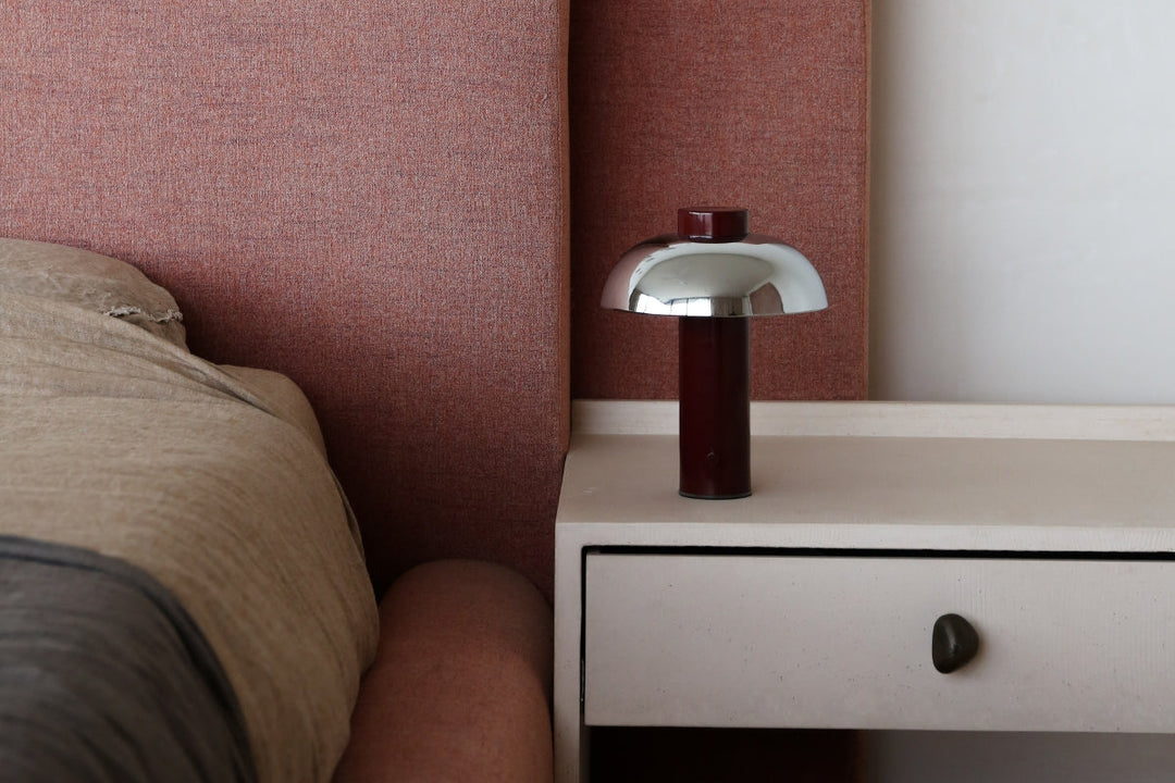 Portable table lamp with chrome dome shade on a bedside table against a warm-toned upholstered headboard.