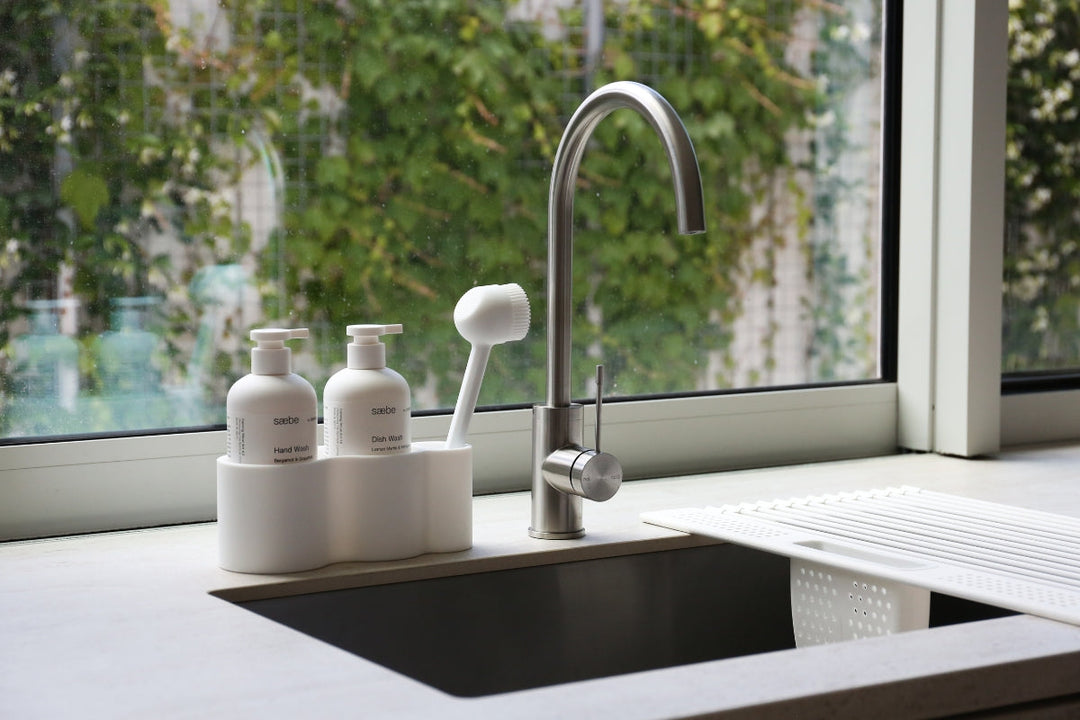 Modern kitchen sink with stainless steel tap, soap dispensers and dish brush in a white sink organiser beside a window.