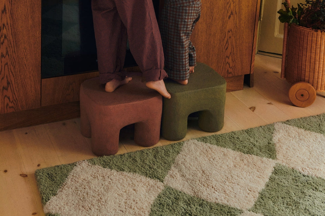 Two children standing on rust and olive sculptural stools on a green-and-cream patterned rug in a timber kitchen.