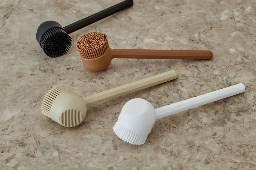VANA silicone dish brushes by DESIGNSTUFF in black, camel, sand, and white, showcasing the seamless silicone construction, ergonomic flat handle, and integrated scraper edge.