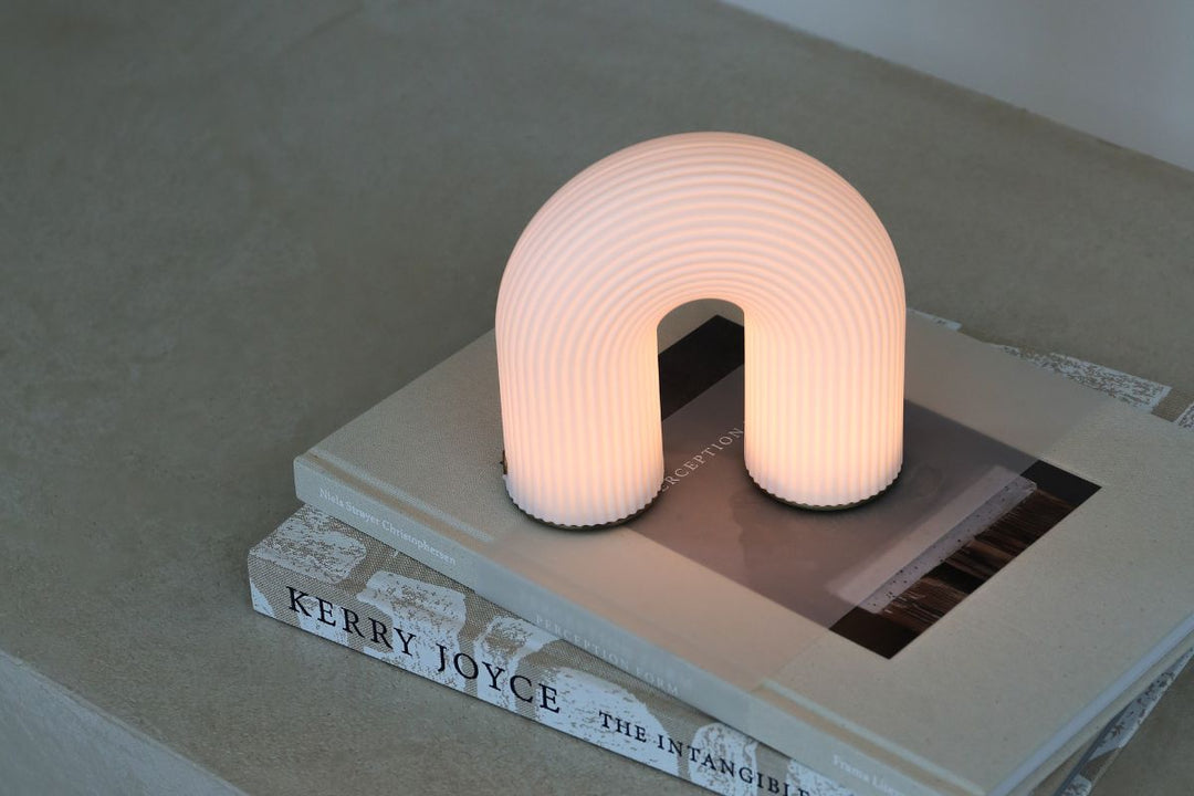 "The Ferm Living Vuelta Portable Lamp emits a soft glow while placed on stacked design books, showcasing its sculptural, modern aesthetic. Discover the best portable lamps for a stylish, minimalist home.