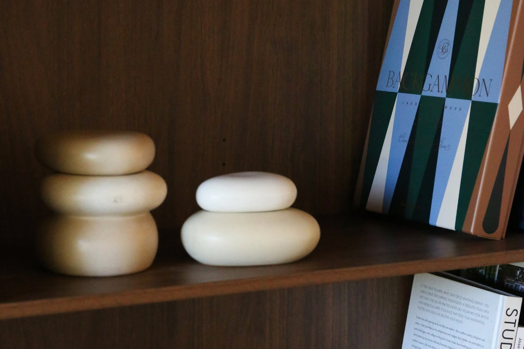 A close-up of a stylishly arranged dark wood shelf displaying two decor pieces with stacked, smooth shapes, alongside a backgammon game box with a blue and green geometric cover.