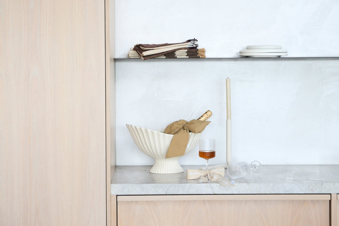 A clean, minimalist composition on a grey stone countertop. A large, fluted ceramic bowl holds a bottle of wine wrapped in paper. Beside it stand a glass of amber liquid and a tall, thin candle. In front, an empty wine glass is tipped over. Above, a float