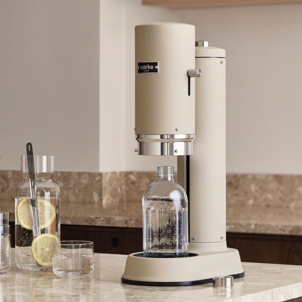 Natural lighting, perspective view of a cannister-shaped carbonator with a glass bottle filled with fizzy water placed on kitchen counter top.