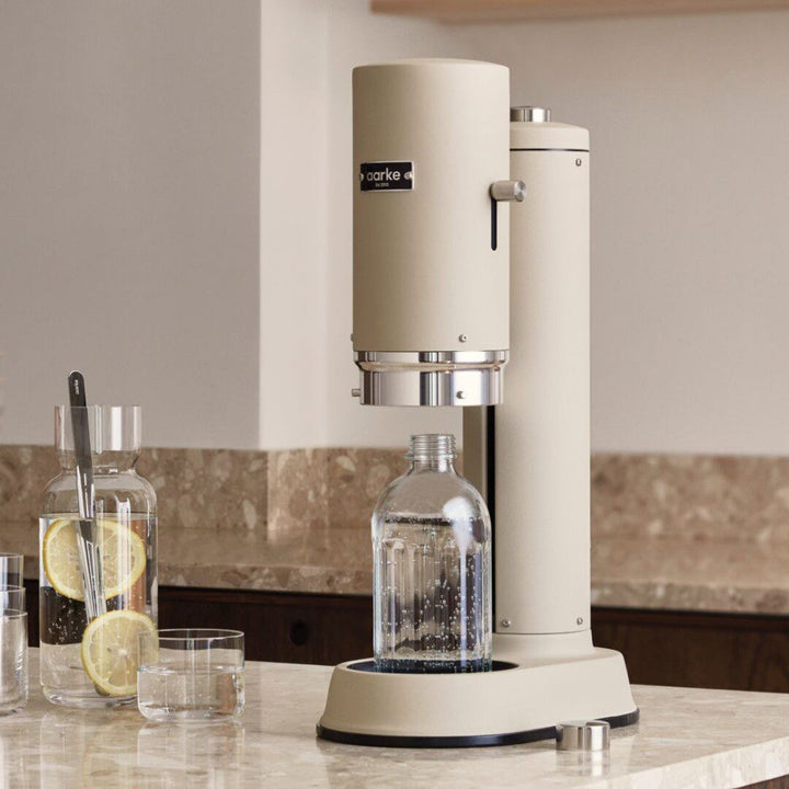 Natural lighting, perspective view of a cannister-shaped carbonator with a glass bottle filled with fizzy water placed on kitchen counter top.