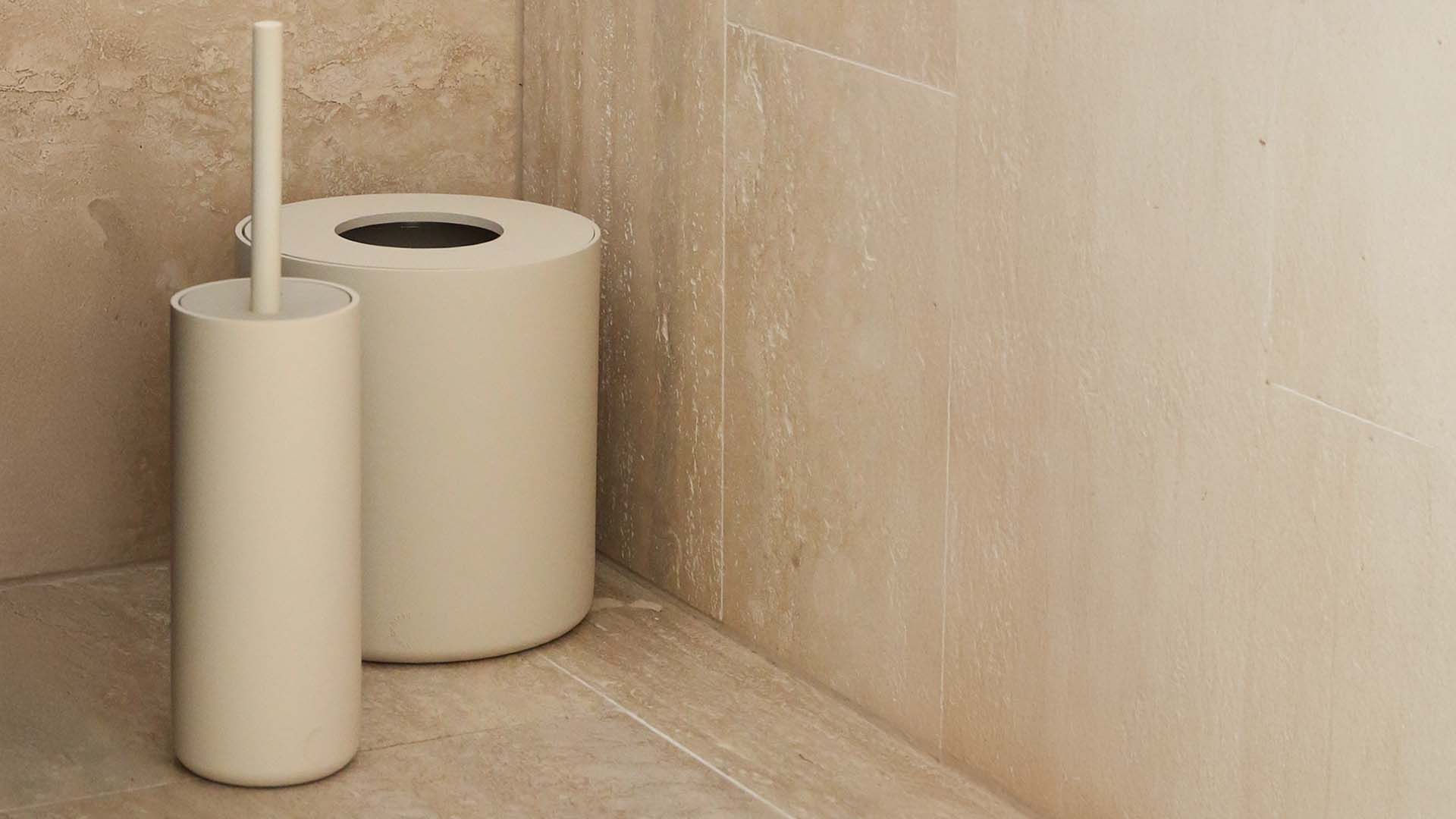 Beige toilet brush holder and roll of toilet paper against a beige tiled wall.