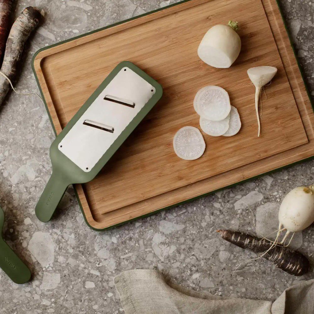 Top view of a kitchen counter with a grater slicer tool producing thinly-sliced radish placed on a chopping board.