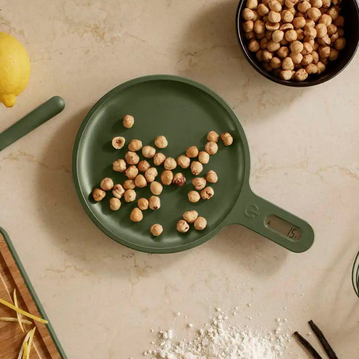 A handful of tree nuts being weighed on a pan-shaped kitchen scale with its weight displayed on the scale's handle.