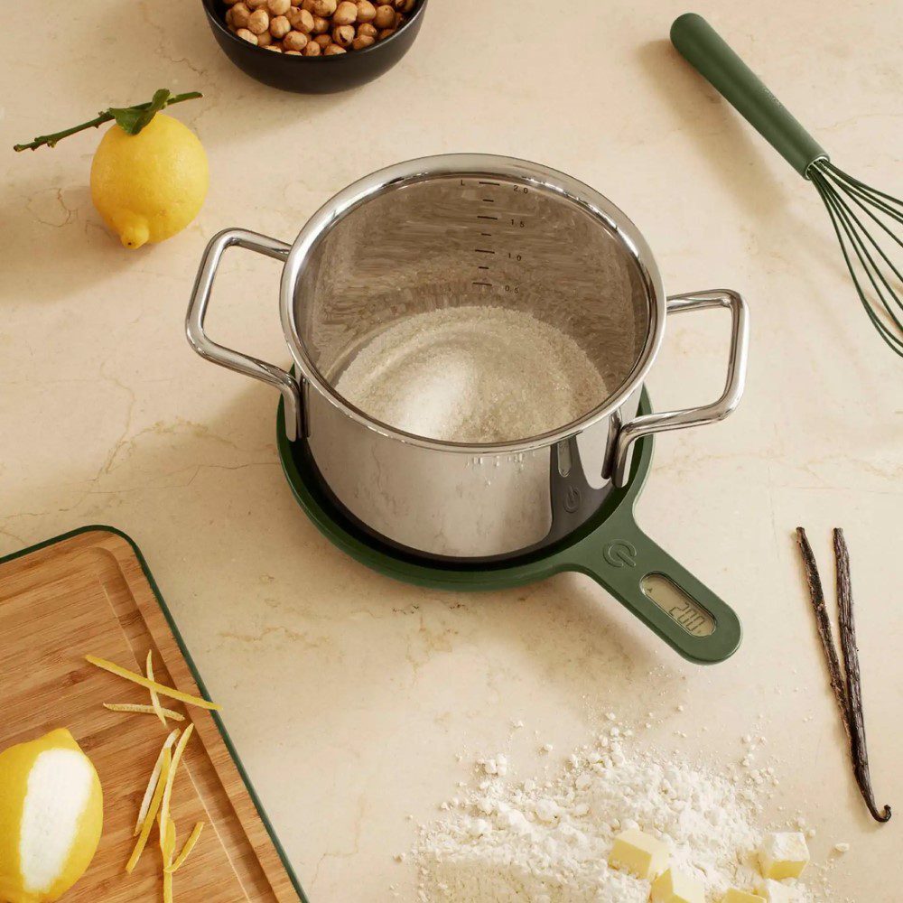 A cooking pot weighed on a pan-shaped kitchen scale with its weight displayed on its handle and placed on a kitchen counter with other utensils.