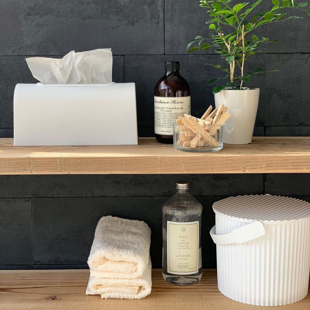 Natural lighting, perspective view of a shelf with a tissue box, bottle of soap, a small pot of flowers, small towels, and a small, cylinder-shaped white cleaning bin placed at the bottom.