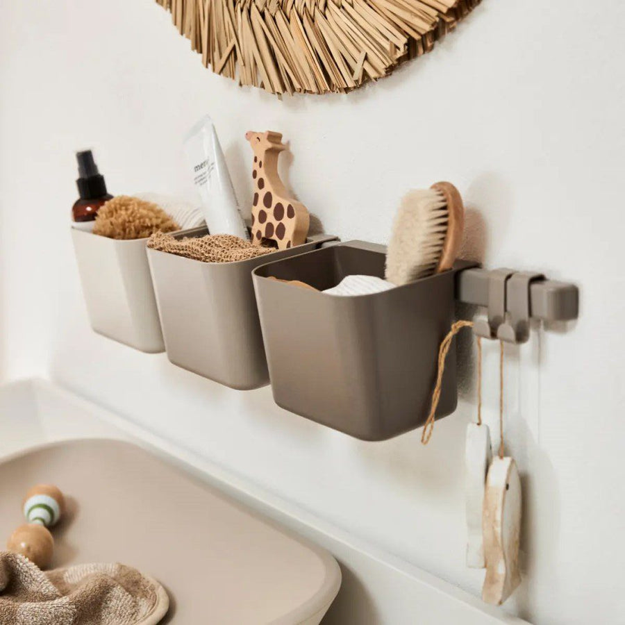 Natural light, close-up image of a wall-mounted, 3-piece grey organiser filled with toys next to a cropped image of a changing pad.