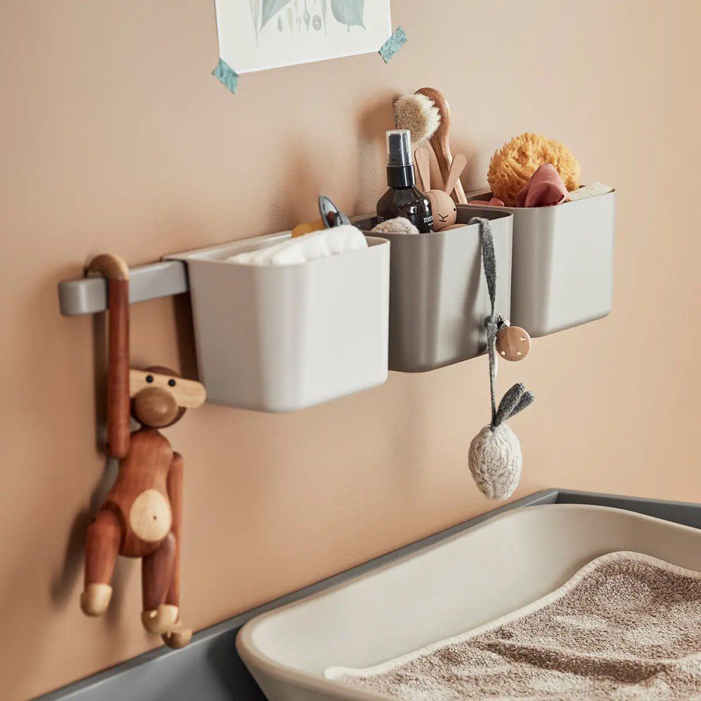 Natural light, close-up image of a wall-mounted, 3-piece grey organiser filled with toys next to a cropped image of a changing pad.