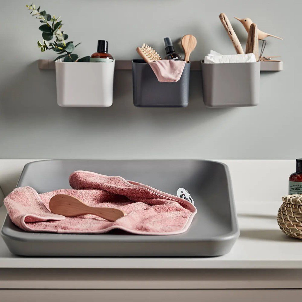 Natural light, perspective view of a grey, 3-piece organiser filled with objects, mounted above silicone changing pad with a pink towel on it.