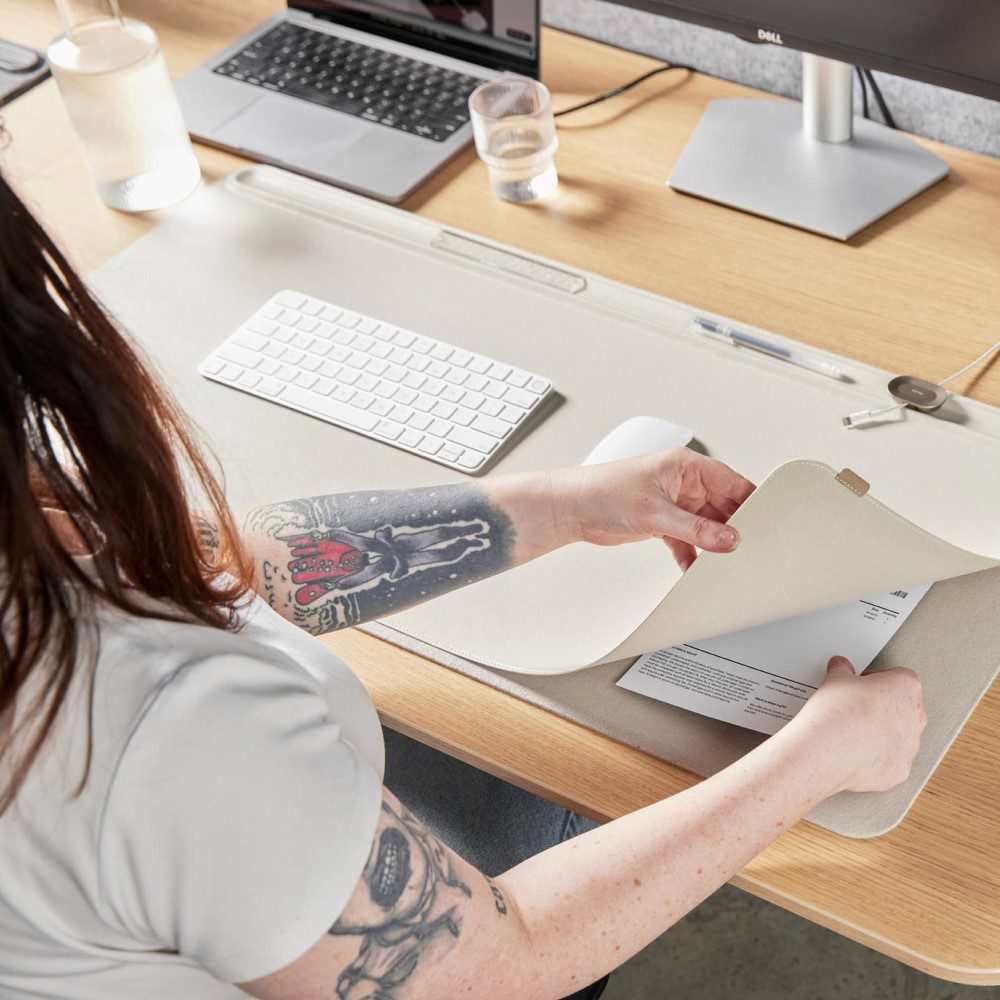 Natural lighting, over the shoulder view of a person placing a written note beneath a rectangular-shaped, light grey desk mat.