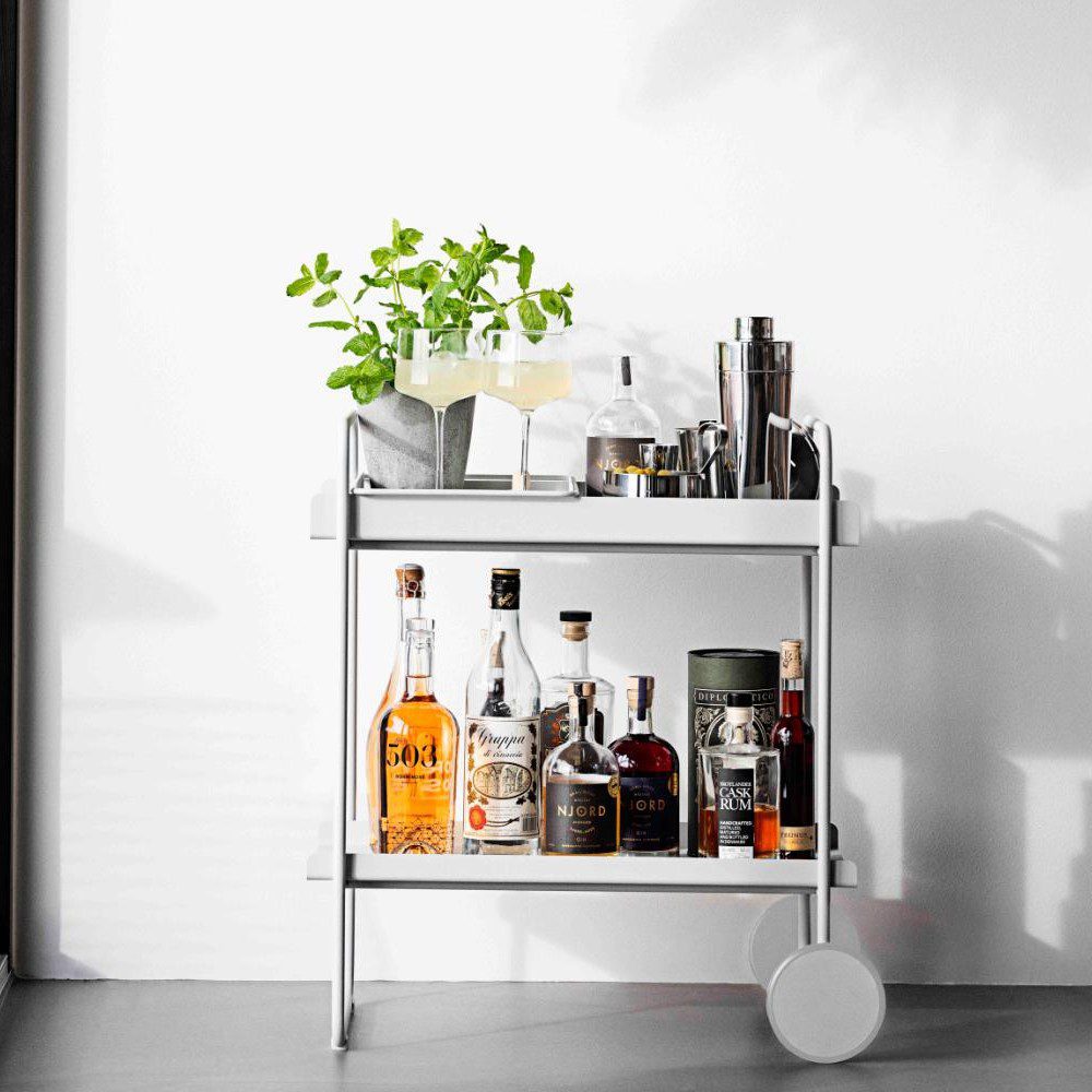 Perspective side view of a cocktail trolley filled with alcoholic drinks, a pot with basil, two cocktail glasses, serving bowls, and a drink shaker.