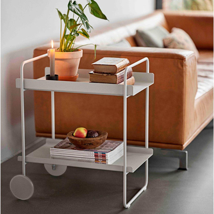 Sun-lit, perspective view of a two-wheeled and tiered cocktail cart and trolley topped with a pot, books, and a display tray.