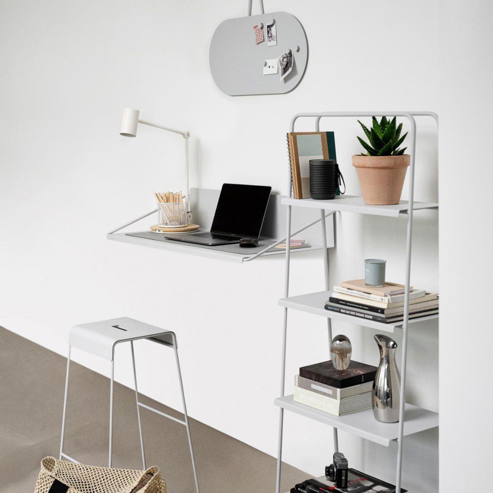 Perspective view of a grey, wall-mounted desk with a laptop beside a four-tier shelf decorated with pots, books,, frames, and objects.