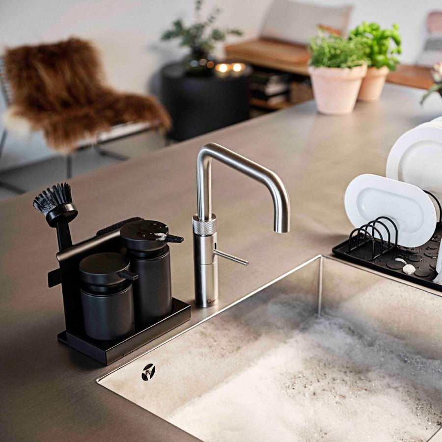 Natural light, isometric view of a stainless steel kitchen sink with a black-coloured tray of cleaning tools beside the sink's faucet.