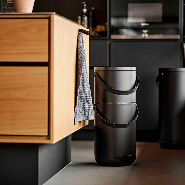 Sun-lit kitchen scene with an image of a tall, black, cylindrical trash bin with a pail-type handle on the floor.