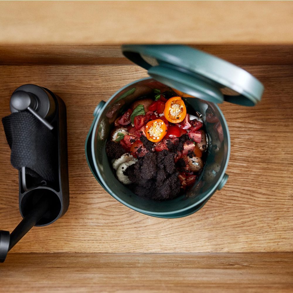 Top view of a small, open, black coloured waste bin containing discarded fruits and vegetables next to a black coloured cleaning kit.