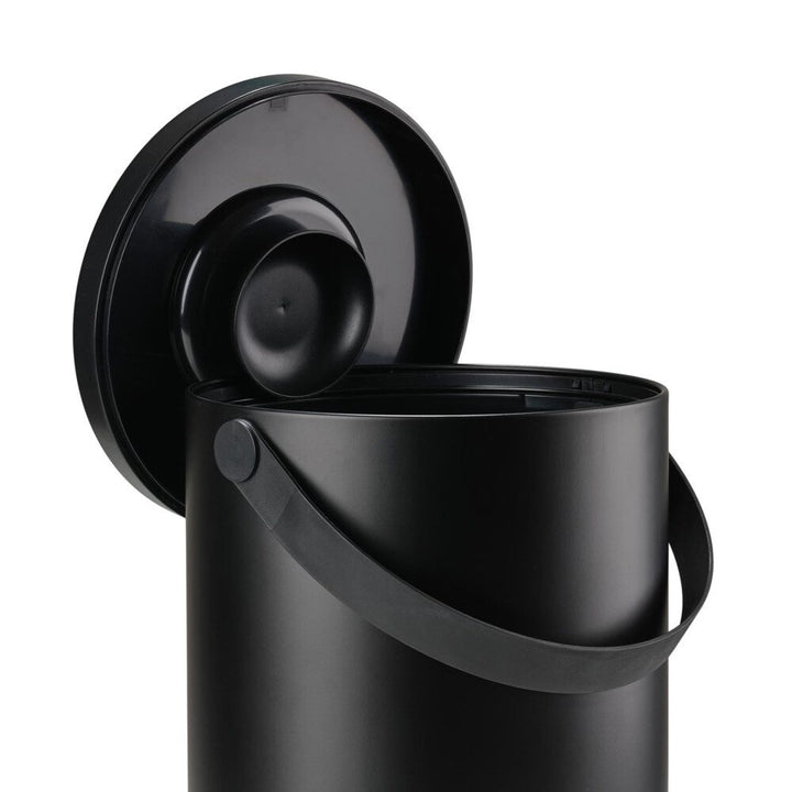 White background, studio image of a black, cylindrical trash bin with a pail-type handle with its cover perched on the rim of the bin.