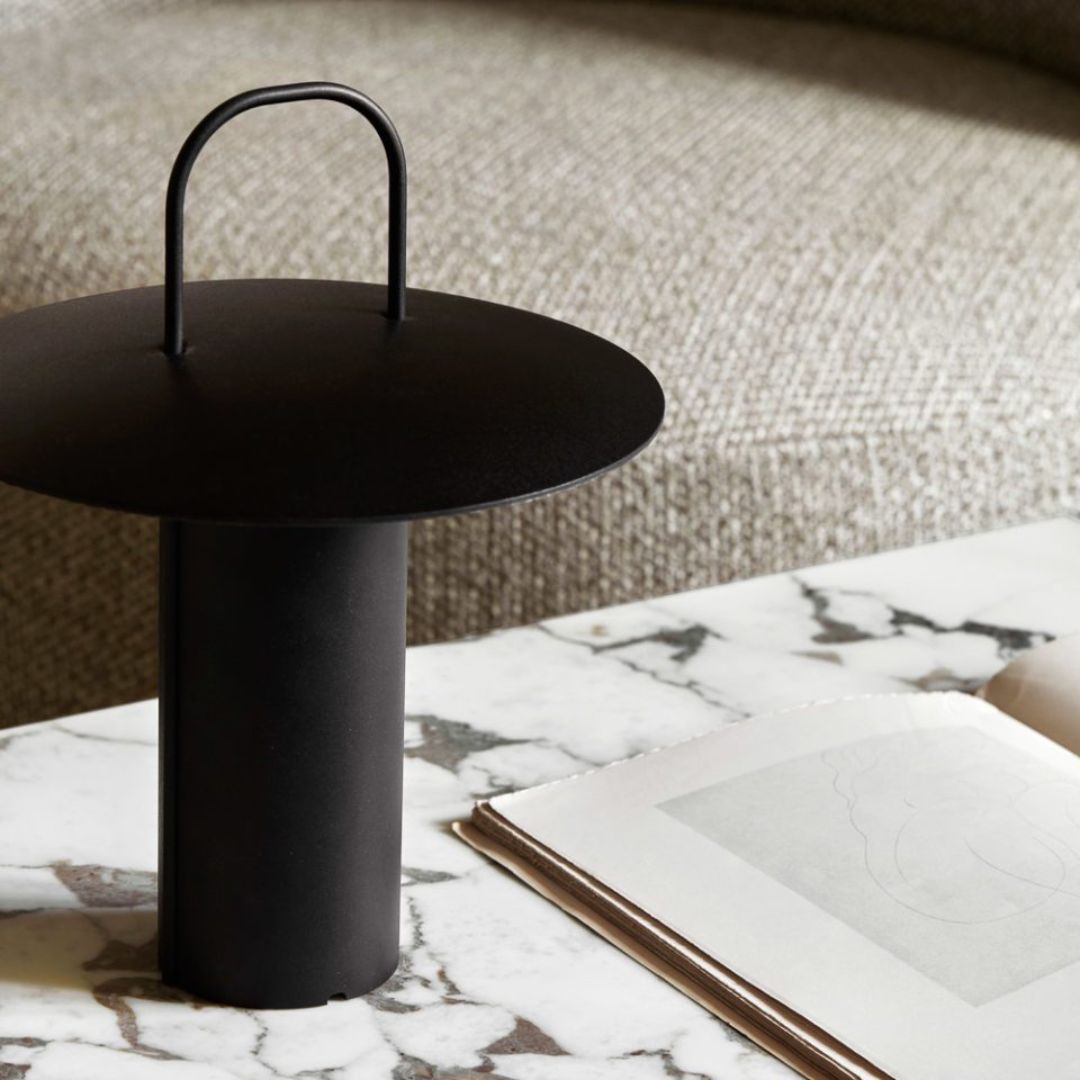 Image features the Menu RAY Table lamp, a portable lamp in black that looks like an upside down top hat. It's sat on top of a veined marble coffee table, alongside a book.